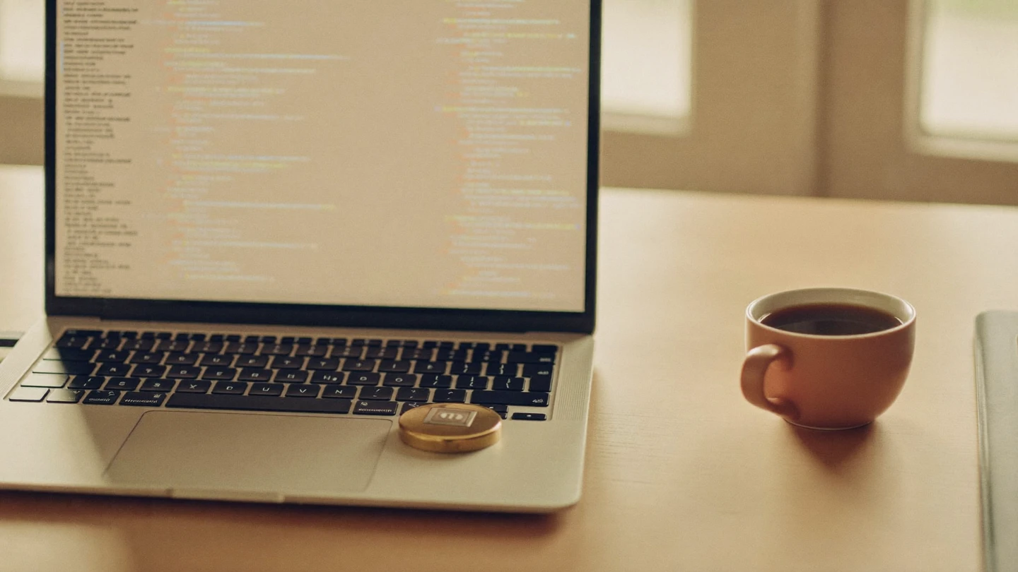 A developer laptop showing blurred code on a warm sunlit desk, with a small brass padlock resting on the keyboard beside a cup of coffee